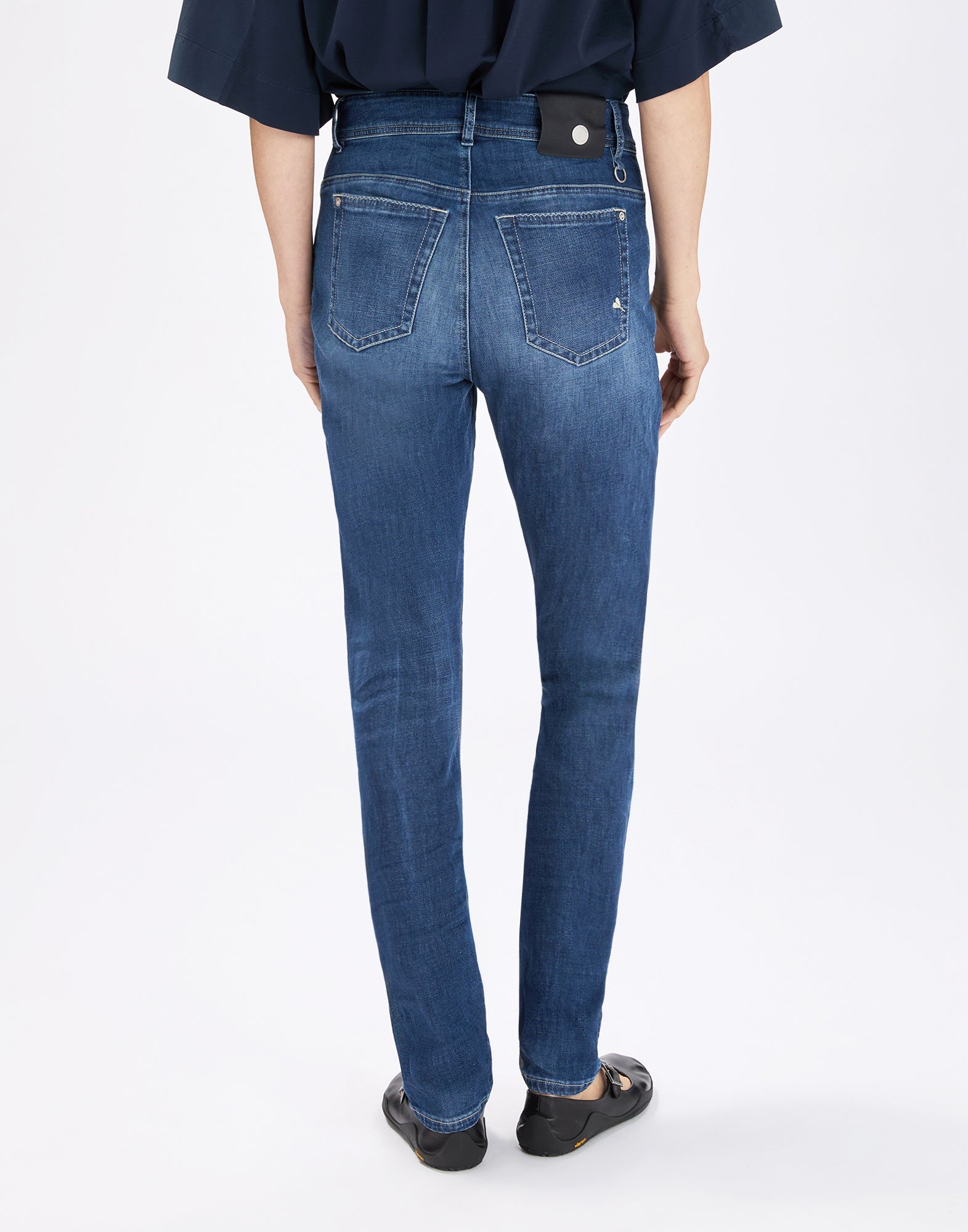 Rear view: Person trägt die enge Jeans OUR-GIRLS in mittlerem Indigo-Blau mit sichtbaren Nähten, zwei aufgesetzten Gesäßtaschen, Gürtelschlaufen und geradem Bein, kombiniert mit dunklem Oberteil.
