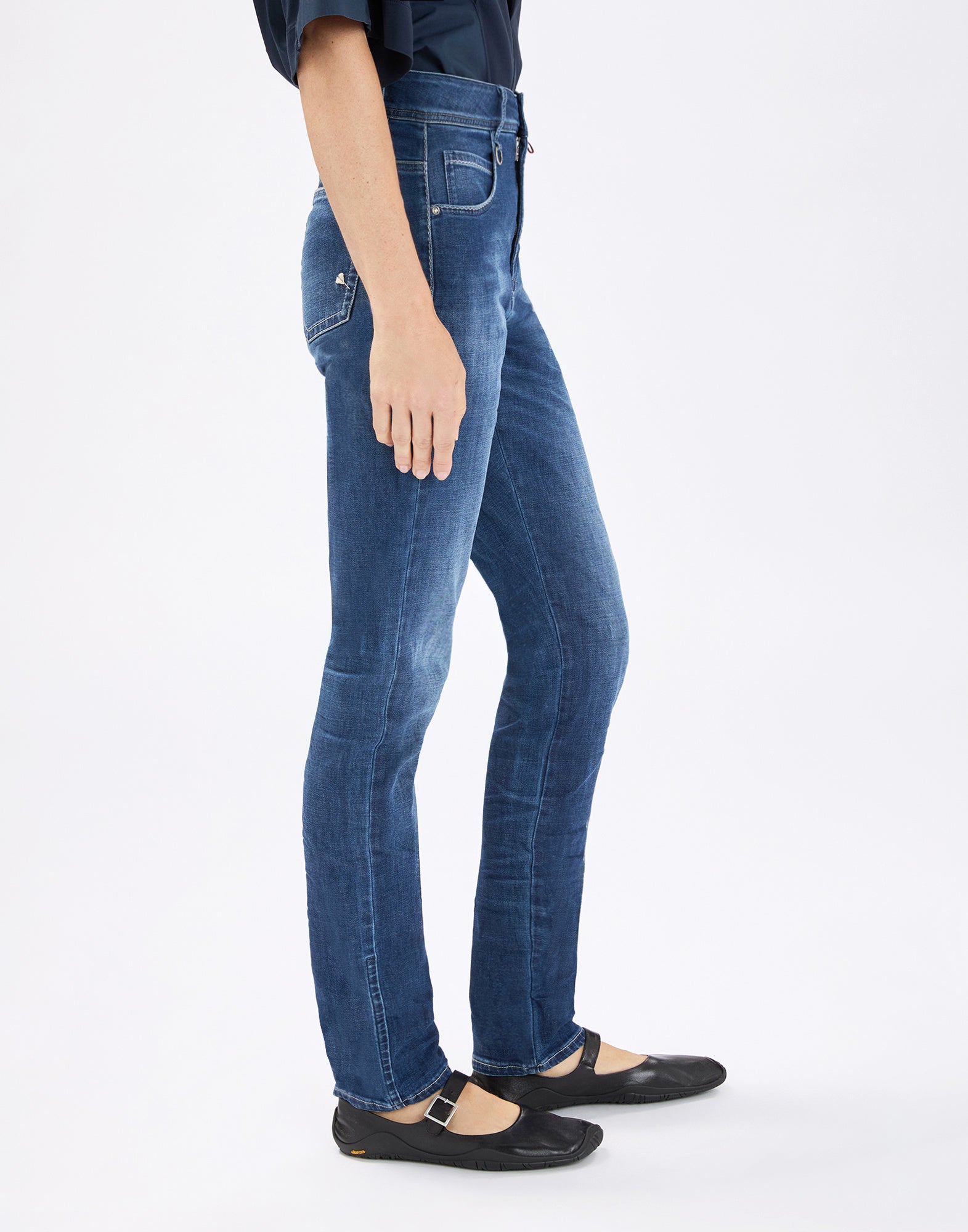 Seitenansicht: Person trägt die eng sitzende Jeans OUR-GIRLS in mittelblauem, leicht verwaschenem Denim mit sichtbaren Nähten, Fronttaschen und Gürtelschlaufen, kombiniert mit schwarzen Mary-Jane-Schuhen.