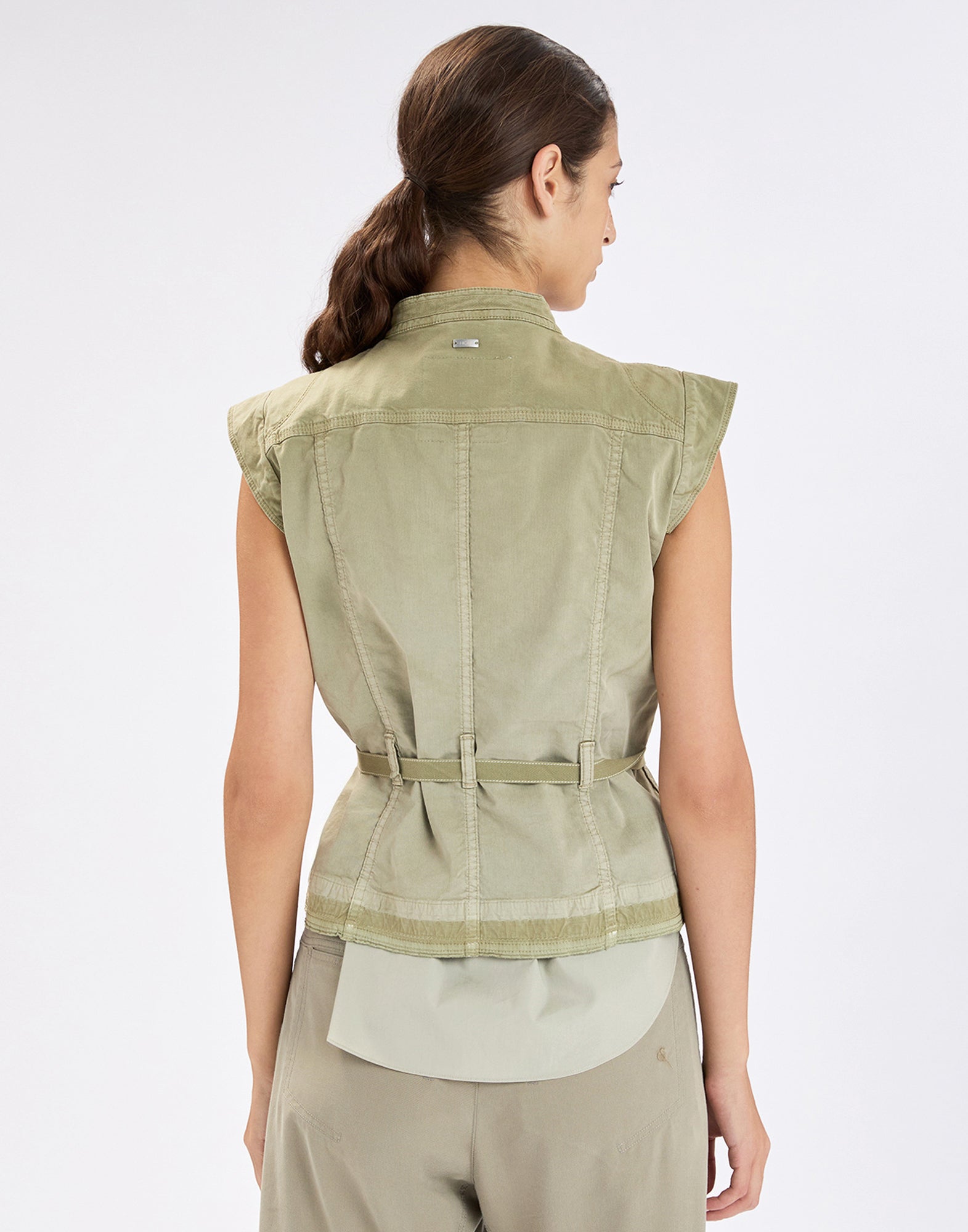Back view: woman wearing the sleeveless stretch gabardine RAISE UP vest in light khaki, with waist belt, visible stitching and shoulder yoke, paired with relaxed-fit trousers.