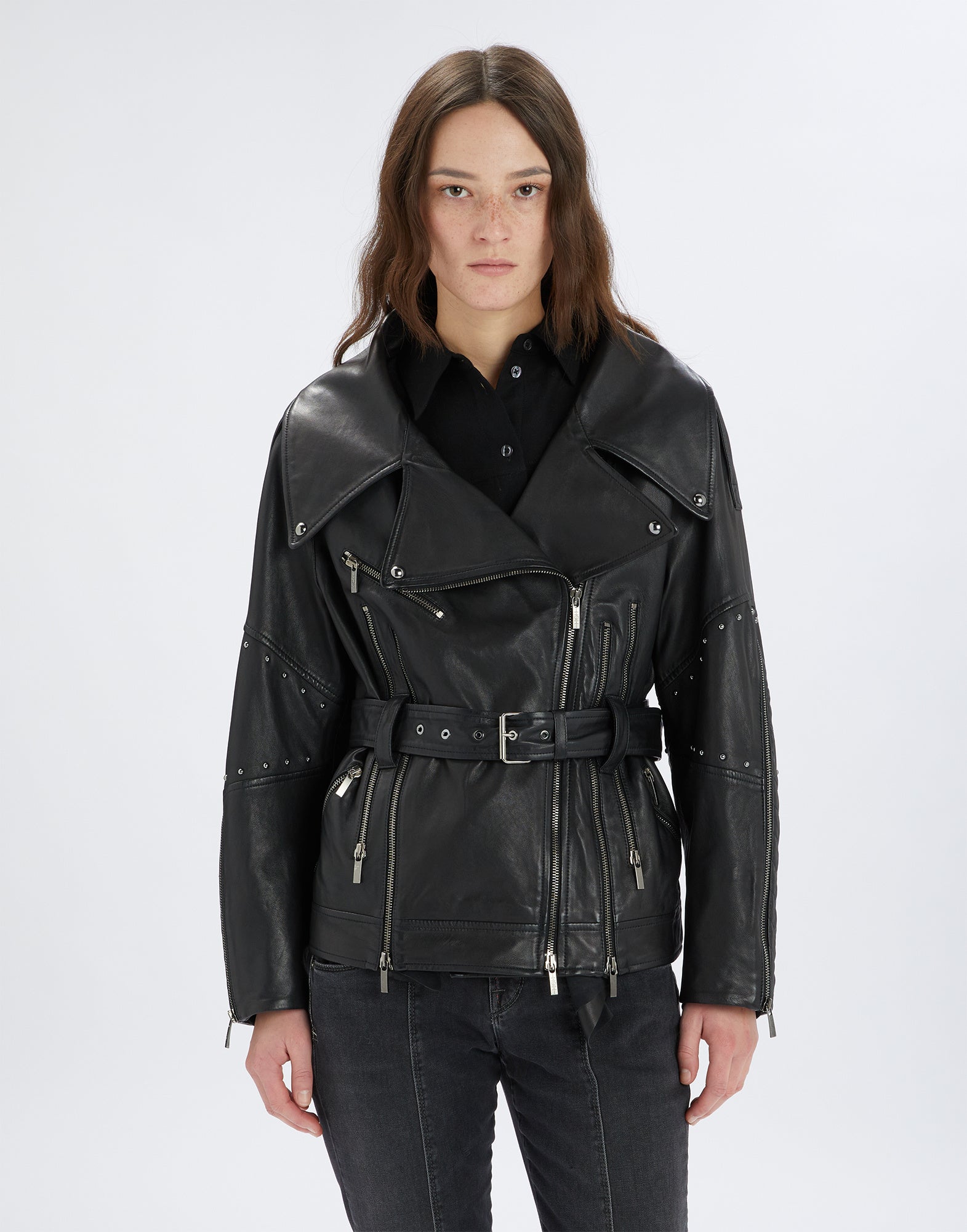 Front view of the TOMMY jacket, black biker jacket featuring prominent notched lapel, multiple zippers, micro studs, and metal buckle belt, hip length.