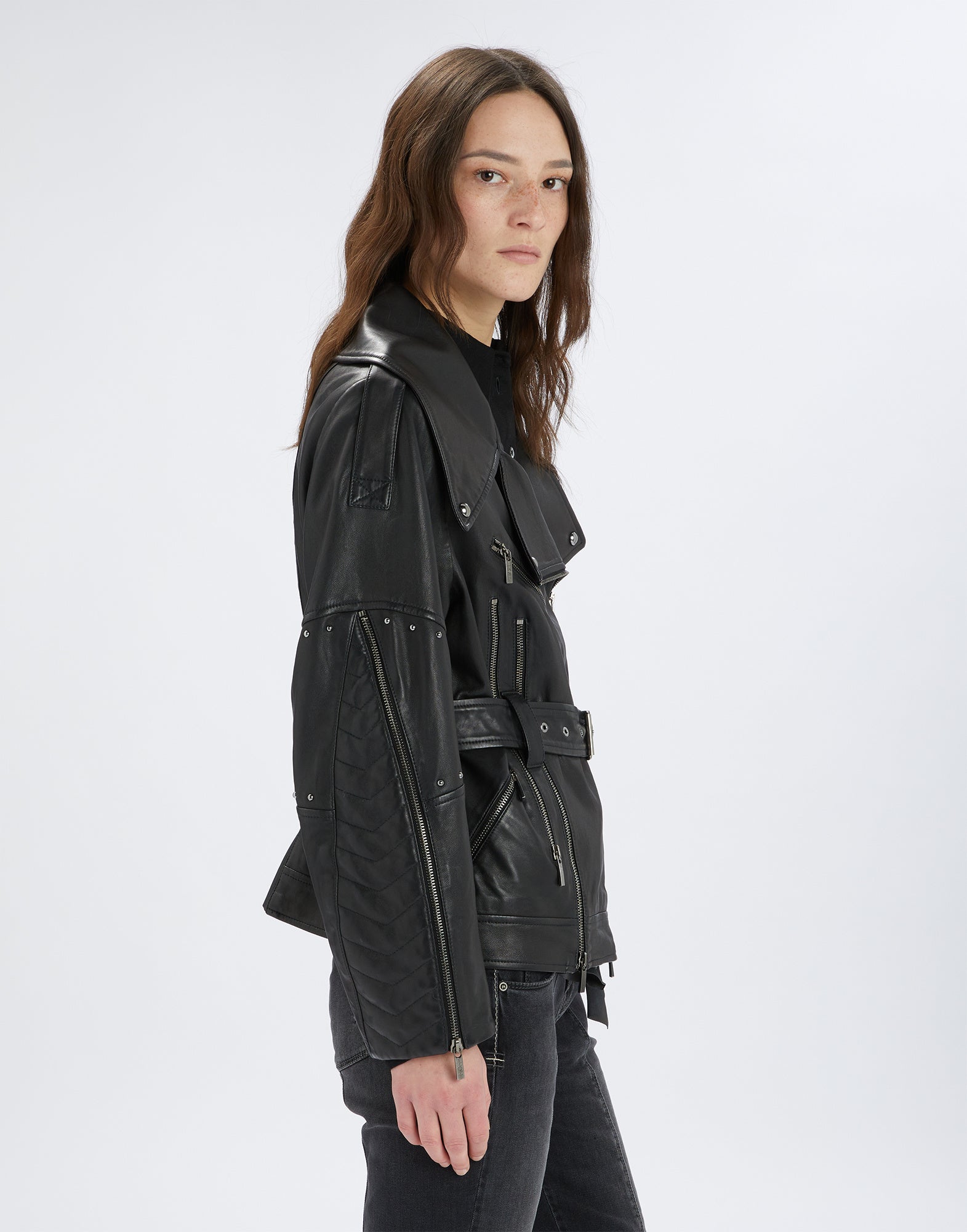 Side view of the TOMMY jacket worn by a woman. Black leather with structured, quilted sleeves, micro studs, zippers, and metal-buckle belt.