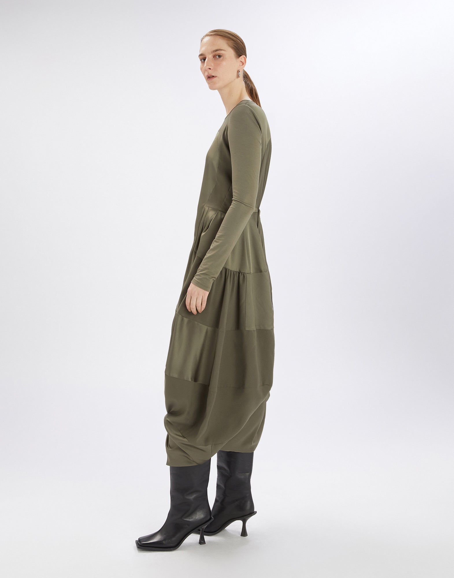 Side view of the PLACID dress, shown on a model. The dress features long sleeves and a flowing olive green satin fabric on the lower part.