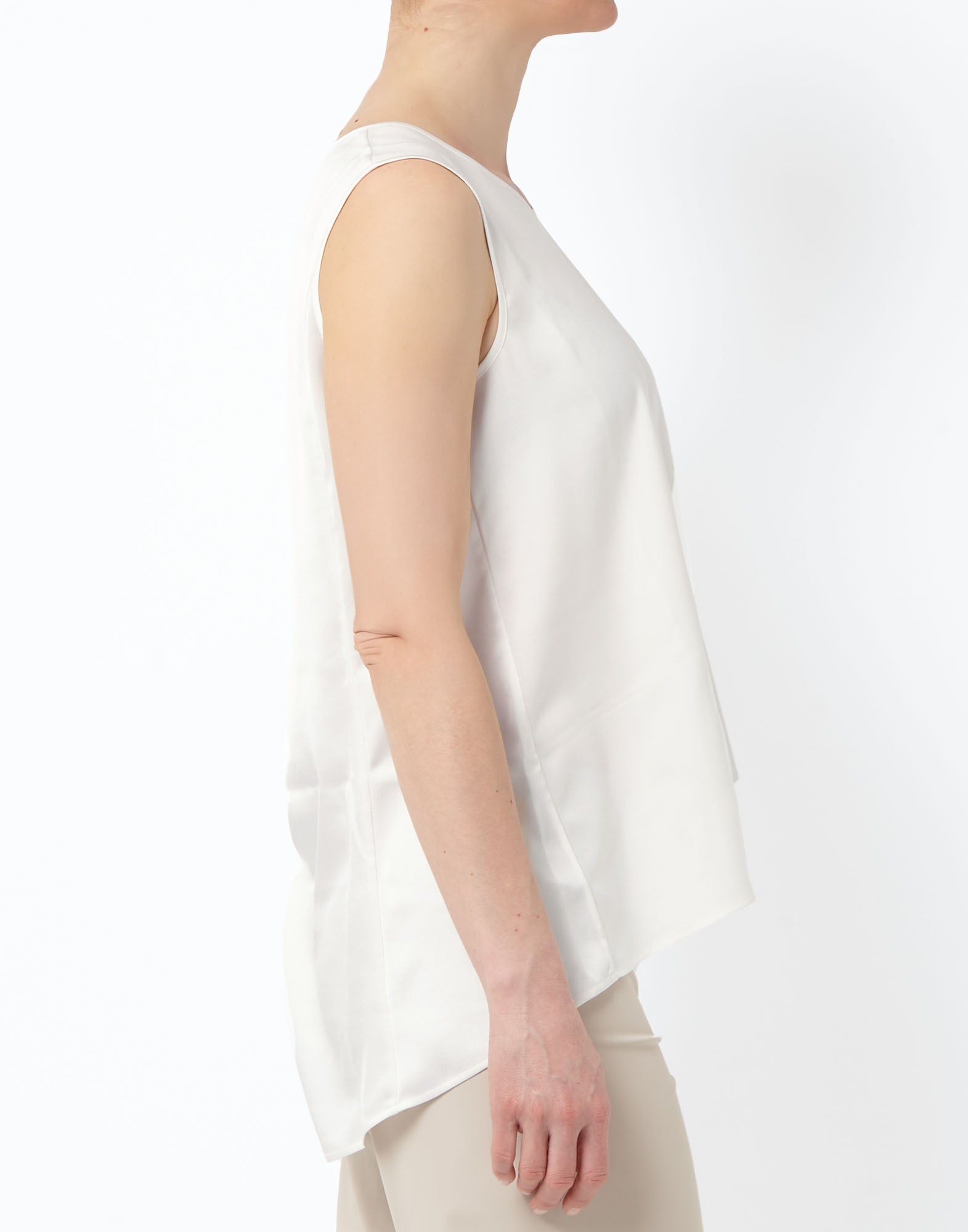 Side view: Model wearing the sleeveless CUE top in slightly shiny technical satin in off-white, with a round neckline and a longer, fluid back, paired with light-colored trousers.