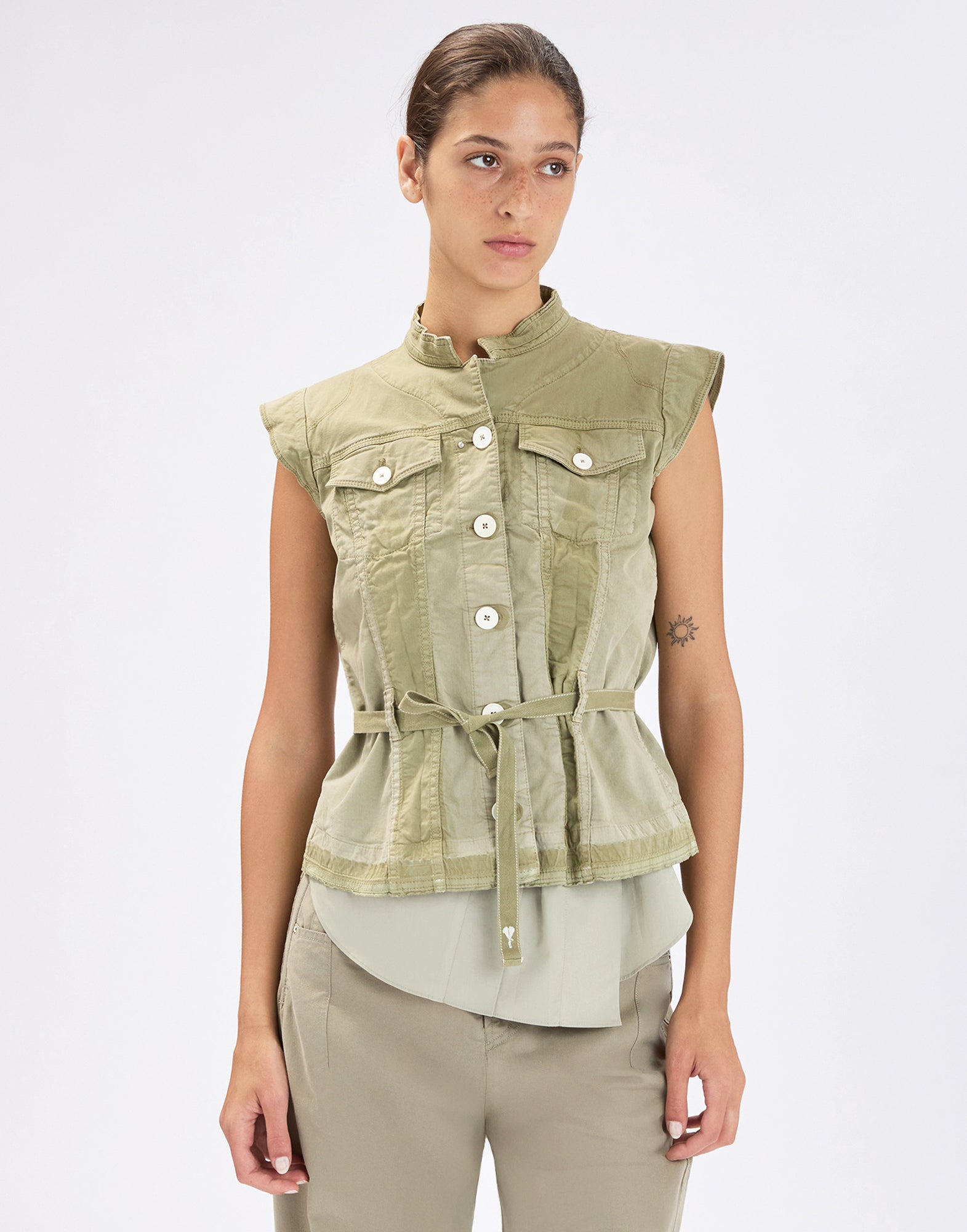 Front view: model wearing the sleeveless, olive green RAISE UP denim jacket with stand collar, front button placket, flap chest pockets and fabric belt at the waist over a light blouse.