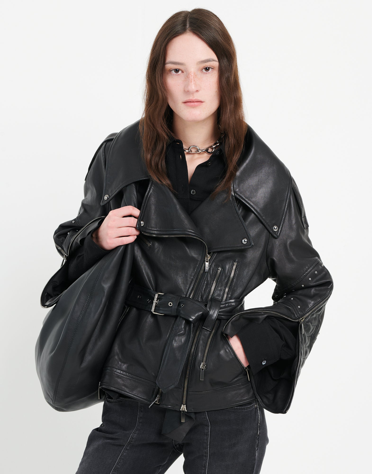 Front view: A woman wears the TOMMY jacket with double-breasted zip, large lapels, metal-buckle belt, and micro studs. Multiple zips and smooth black leather define the design.