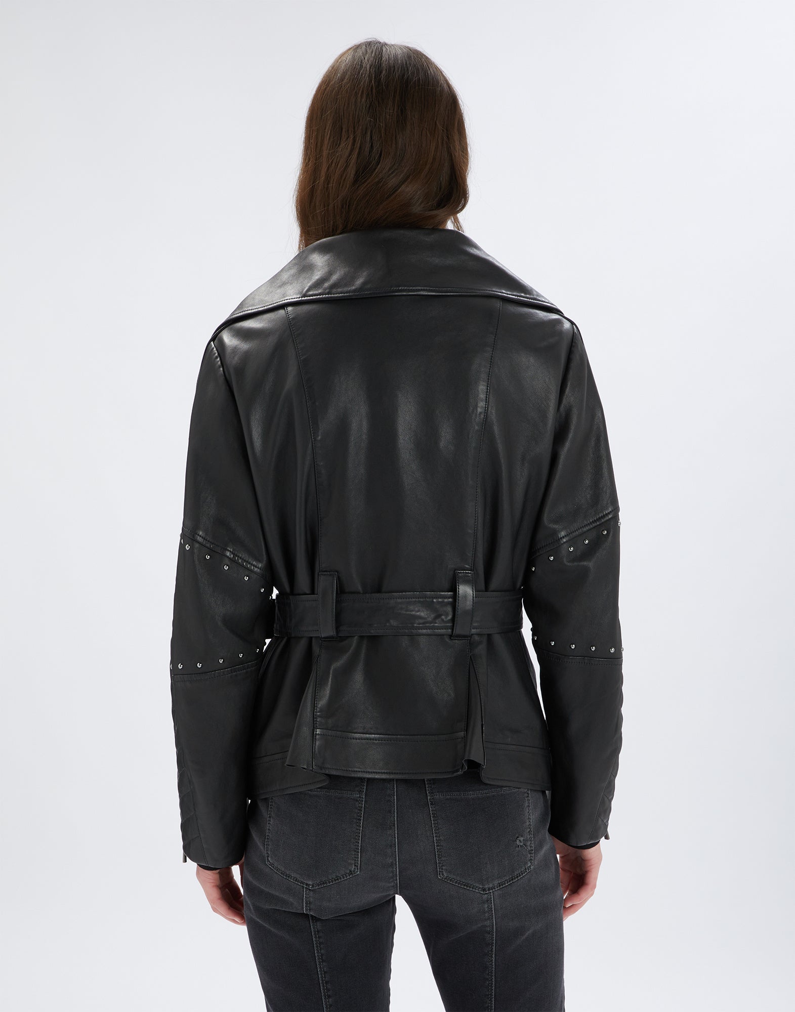 Rear view of the TOMMY jacket, worn by a person. Black leather with micro studs on the sleeves, waist belt, and classic collar visible.